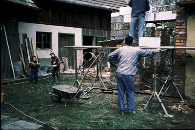 Four construction workers in a courtyard, mid-1980s Eastern Bloc. Two men on scaffolding install a metal beam; one on ground ...