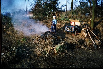 A person in vintage outdoor workwear stokes a compost pile with smoke rising, beside a small tractor with a hay rack. Rural s...