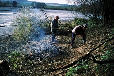 Two men clear brush by a riverbank, likely for agricultural or flood prevention. One kneels, using a tool to cut vegetation; ...