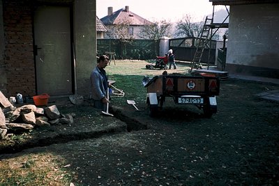 Mid-20th century backyard scene featuring two men in work attire. One man stands near a brick garage door holding a shovel, w...