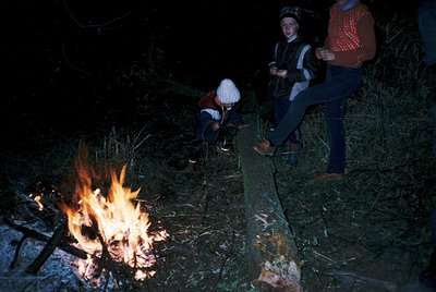 Vintage nighttime campfire scene with three individuals in 1970s outdoor attire—striped sweaters, jeans, and headscarves. One...