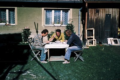Three men seated at a foldable table in a backyard, likely mid-1970s. Light green house with white-framed windows and a woode...