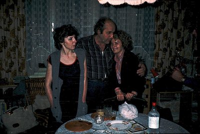Family gathering in dimly lit room with patterned wallpaper, likely 1970s–1980s. Three adults—man in plaid shirt, woman in bl...
