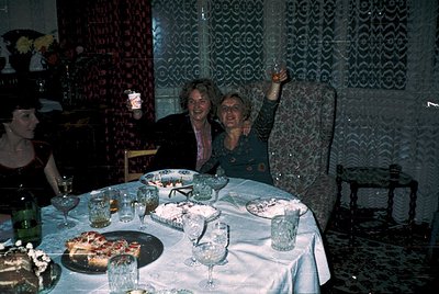 Vintage indoor celebration with three women at a round table adorned with cake, glasses, and floral centerpieces. Decor inclu...