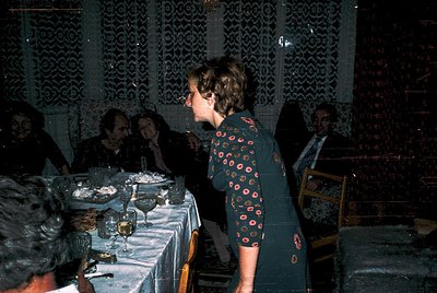 Vintage indoor gathering in a dimly lit room with patterned wallpaper, featuring a woman in a floral-patterned dress serving ...