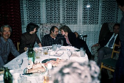 Five men gather around a long table in a dimly lit room with patterned lace curtains, likely a 1970s-80s social gathering. Ta...