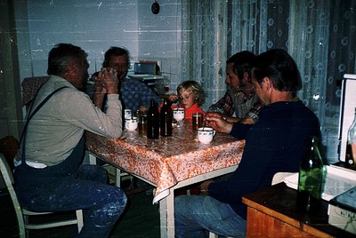 Four adults and a child gather around a round table in a dimly lit room, likely a 1970s-era home or café. The table features ...