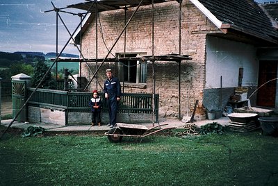 Rural brick home with exposed scaffolding, likely mid-20th century. Two men in dark uniforms (one adult, one child) pose on a...