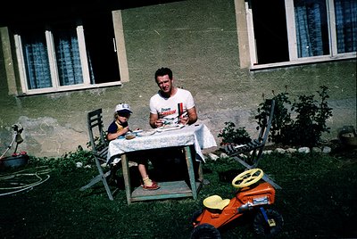 A man and child seated at a foldable metal table outdoors, likely 1970s Eastern Europe. The man wears a white T-shirt with re...