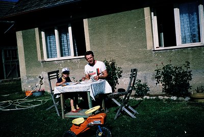 Mid-20th century family moment outside a weathered concrete house. Man in white shirt with "Coca-Cola" logo sits at a foldabl...