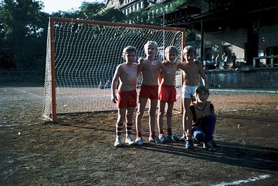 Five children pose in front of a soccer net on a dirt field, mid-1970s. Four stand in matching red shorts with white stripes;...