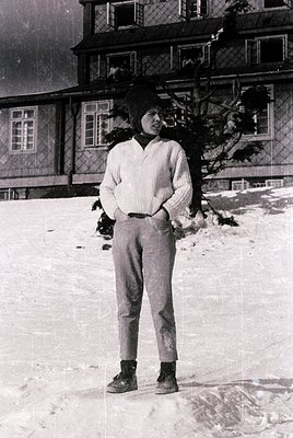 Young individual in 1960s ski attire—V-neck sweater, wide-leg trousers, and boots—standing on snow-covered ground beside a wo...