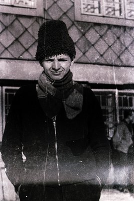 Portrait of a man in winter attire—black fur hat, scarf, and coat—standing in front of patterned brickwork. Likely mid-20th c...