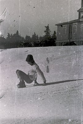 Vintage black-and-white photo of a child skiing on a groomed slope, crouched in a classic stance. Snow-covered pine trees and...