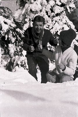 Two men pose joyfully in winter attire amidst snow-covered evergreens, likely mid-20th century. The seated man holds a snow-c...
