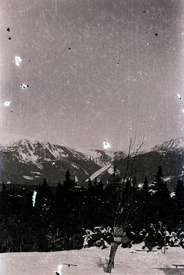 Vintage sepia-toned alpine landscape with snow-covered peaks and dense coniferous forest. Prominent wooden cross and gravesto...