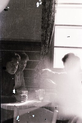 Vintage black-and-white interior shot of three seated figures in a dimly lit room, likely a café or diner. Two women in mid-2...
