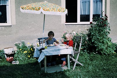 Vintage outdoor picnic scene with two children seated at a wooden table under a floral-patterned umbrella. The setting appear...