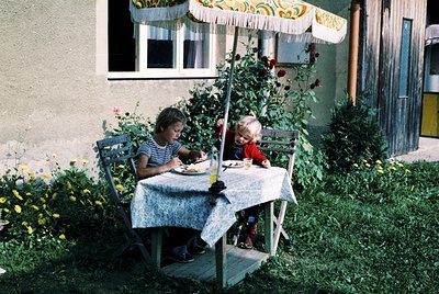 Two children dining al fresco under a striped patio umbrella, seated at a small wooden table with floral tablecloth. The adul...