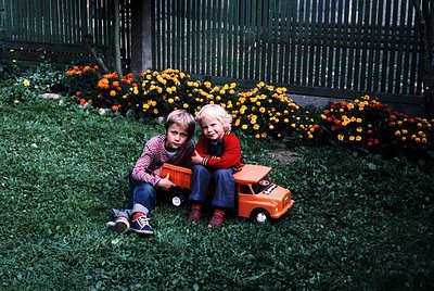 Two children in 1970s-style clothing pose with a toy truck in a vibrant garden bed of orange and yellow chrysanthemums. The b...