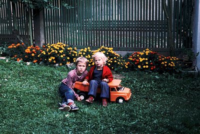 Two children sit on lush green grass beside a vintage toy truck, surrounded by vibrant orange chrysanthemums. The boy in red ...
