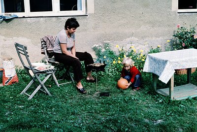 A woman in casual 1970s attire sits on a folding chair beside a child playing with a ball in a grassy backyard. Whitewashed s...