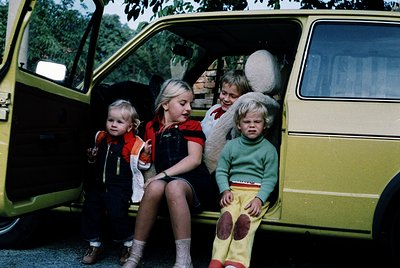 Four children in 1970s-style clothing sit in an open yellow van, likely a VW Transporter. The girl on the left wears a stripe...