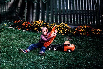 Vintage childhood scene: child in 1970s-style striped shirt and denim shorts plays with a toy truck on lush green lawn. Vibra...