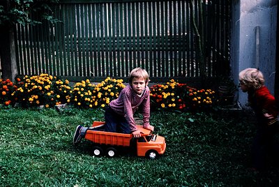Vintage 1970s backyard scene featuring two children playing with a red toy truck on lush green grass. Bright yellow flowers i...