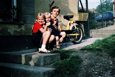 Family portrait on concrete steps, 1970s suburban setting. Three individuals—two children and an adult—pose together; adult h...