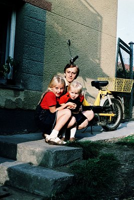 A mid-20th-century family moment: an adult kneels on concrete steps holding two young children (girl in red, boy in yellow) b...