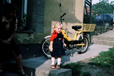 Vintage yellow moped with wire basket parked beside a young girl in a 1970s-style dress and overalls, standing on concrete st...