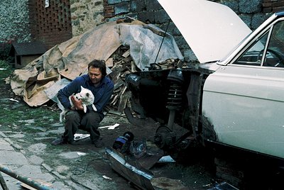 Man in 1970s workwear crouches beside a damaged vintage car, holding a dog. Hood open reveals exposed engine components and d...