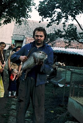 Man in 1970s workwear holds a live beaver, likely for fur trade or farming. Rural setting with wooden houses and a crowd of o...