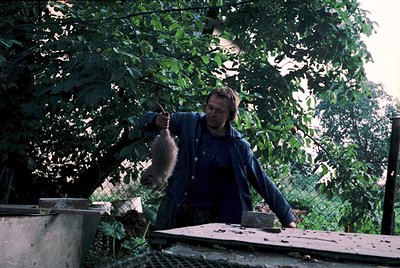 Man in 1970s-style long-sleeve shirt and pants holding a young bear cub outdoors under leafy trees. Wooden platform and concr...