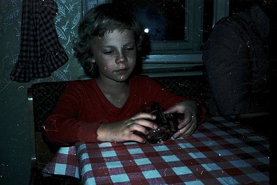 Vintage black-and-white photo of a young child seated at a table with a checkered red-and-white tablecloth, holding a small m...