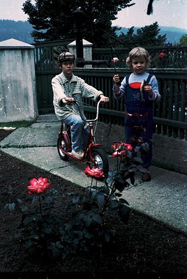 Two children in 1970s-style clothing pose outdoors on a paved area with a wooden fence and greenery. The boy on a red pedal b...