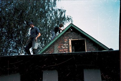 Two men repair a rustic brick house roof, likely mid-20th century. One holds a shovel, the other a bucket, under a green meta...
