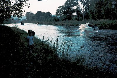 Vintage black-and-white photo of a child standing on a riverbank, observing a motorboat passing. Overgrown vegetation frames ...
