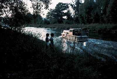 Two children stand on riverbank near a wooden boat, surrounded by dense foliage. The scene evokes mid-20th century rural life...