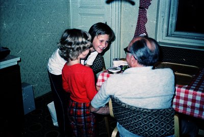 Vintage indoor family moment: three children and an elderly man in a dimly lit room, likely mid-20th century. The man, wearin...