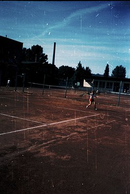 Vintage tennis court with cracked clay surface under clear skies, framed by institutional brick buildings. Player mid-serve, ...
