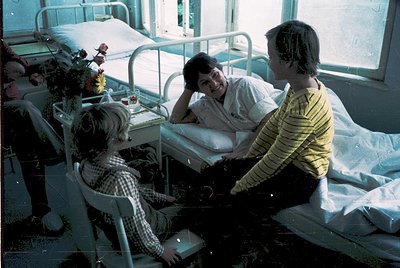1970s hospital ward scene: three individuals—patient in striped hospital gown, nurse in white uniform, and visitor in striped...