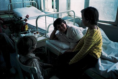 Vintage hospital ward scene from the 1970s–80s, featuring three individuals in a shared room. A nurse in a white uniform sits...
