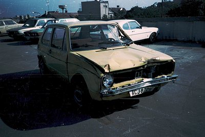 Vintage hatchback with severe front-end damage, likely a 1970s-era Fiat 127 based on design. Parked in a lot with other class...