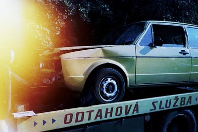 Vintage yellow station wagon lifted on a flatbed truck labeled "ODTAHOVÁ SLUŽBA" (tow service) under golden-hour lighting. Di...