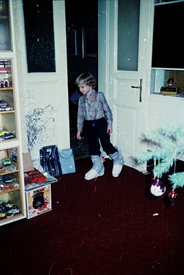 A young child in 1970s-style plaid shirt, dark pants, and white sneakers stands in a dimly lit hallway, framed by white doubl...