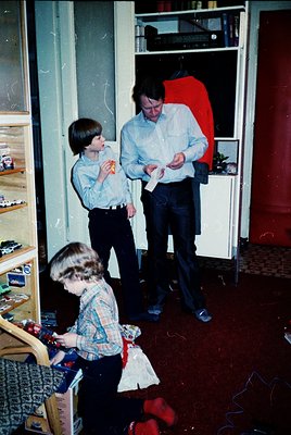 Vintage indoor scene featuring three children in 1970s-style clothing playing in a cluttered room. Two boys in button-up shir...