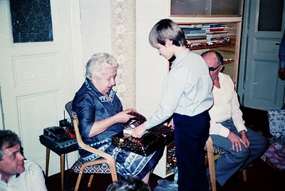 Vintage indoor scene featuring three adults engaged in a craft activity, likely embroidery or beadwork. Elderly woman seated ...