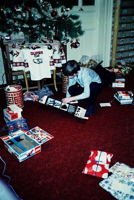 A child kneels on a red patterned carpet, assembling a Lego set near a decorated Christmas tree. Surrounding them are wrapped...